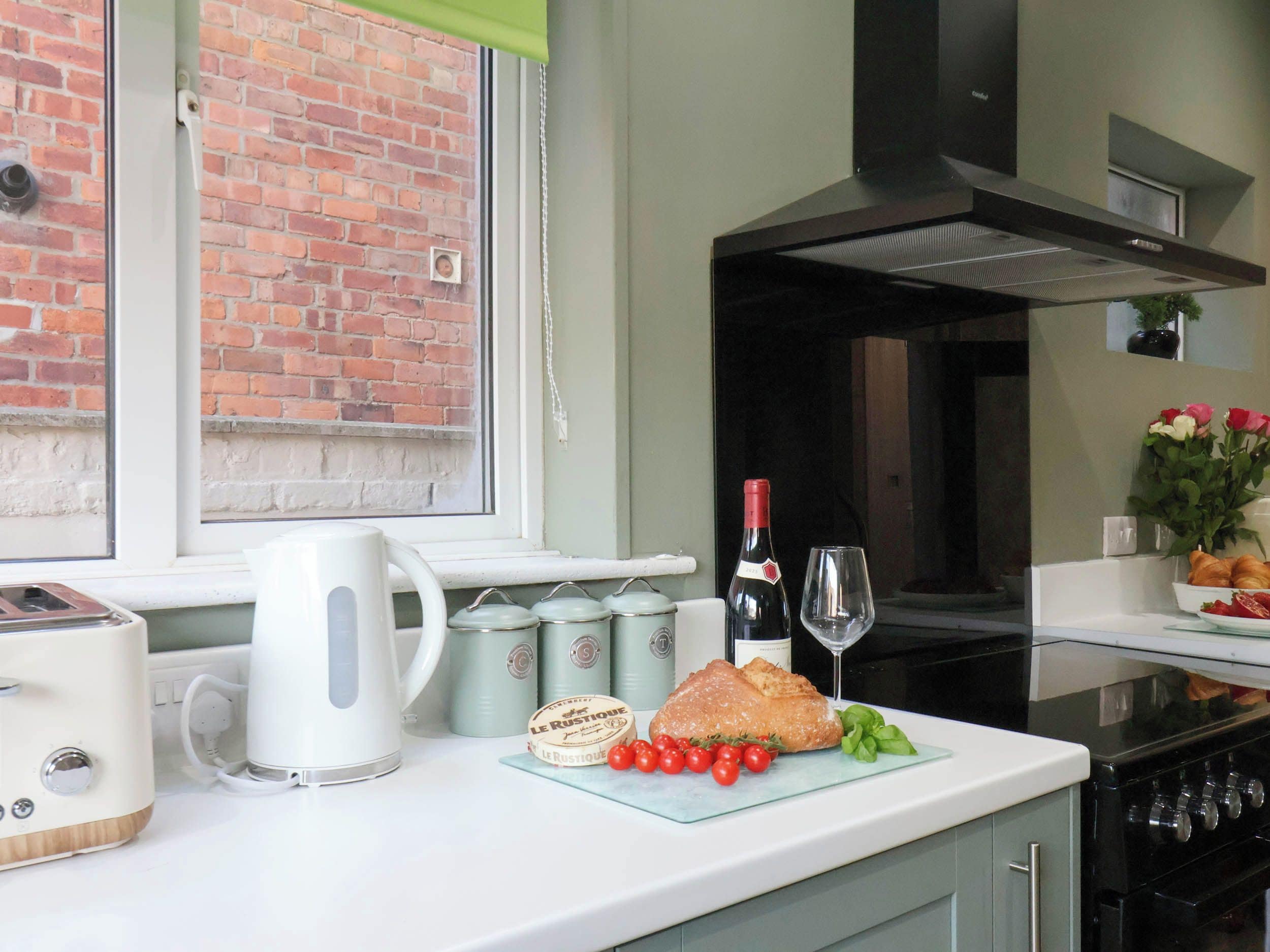 A kitchen with a brick wall, a window, a toaster, a kettle, a wine bottle, a glass, a croissant, cherry tomatoes, and a cutting board.