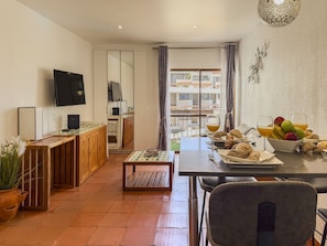 A modern living room with a wooden dining table, a TV mounted on the wall, a balcony with a view of buildings, and a breakfast setup with fruits and pastries. - #409 Central Albufeira Getaway with Shared Pool (Albufeira)