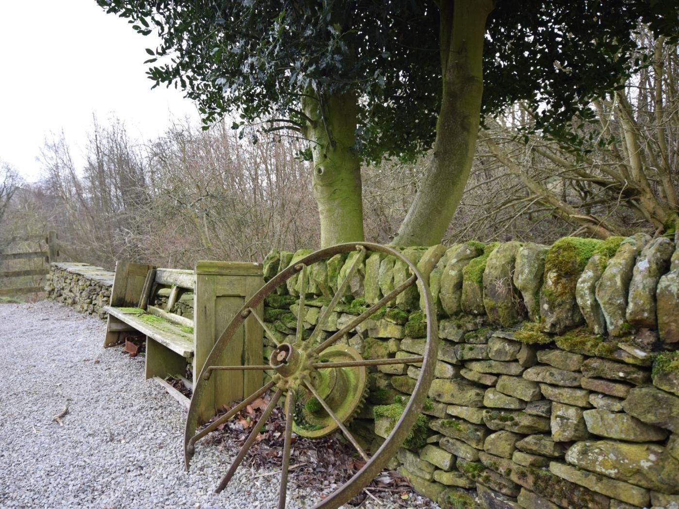 A rustic stone wall with a large wheel, a wooden bench, and a tree in the background.