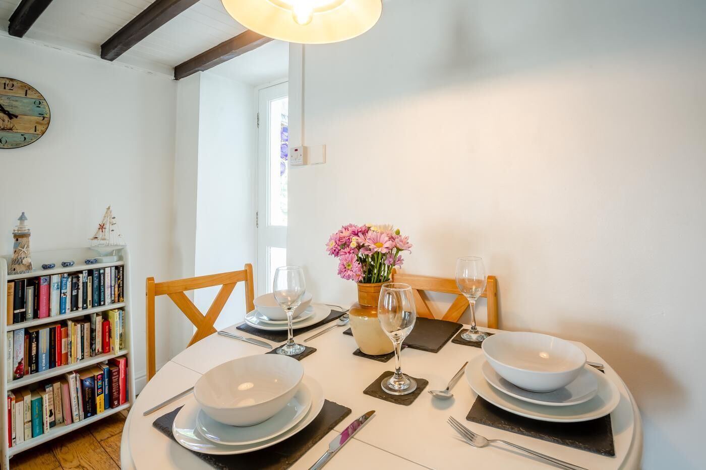 Un comedor con una mesa redonda dispuesta para dos, una estantería con libros, un modelo decorativo de barco y un reloj de pared.