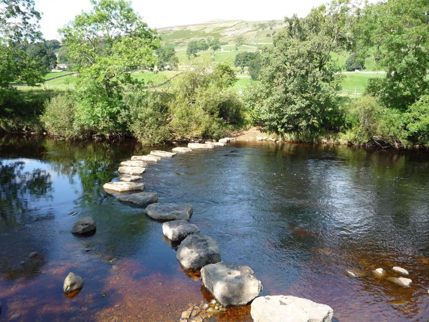 Une rivière traversée par des pierres qui permettent de la traverser à pied, entourée de verdure et de collines.
