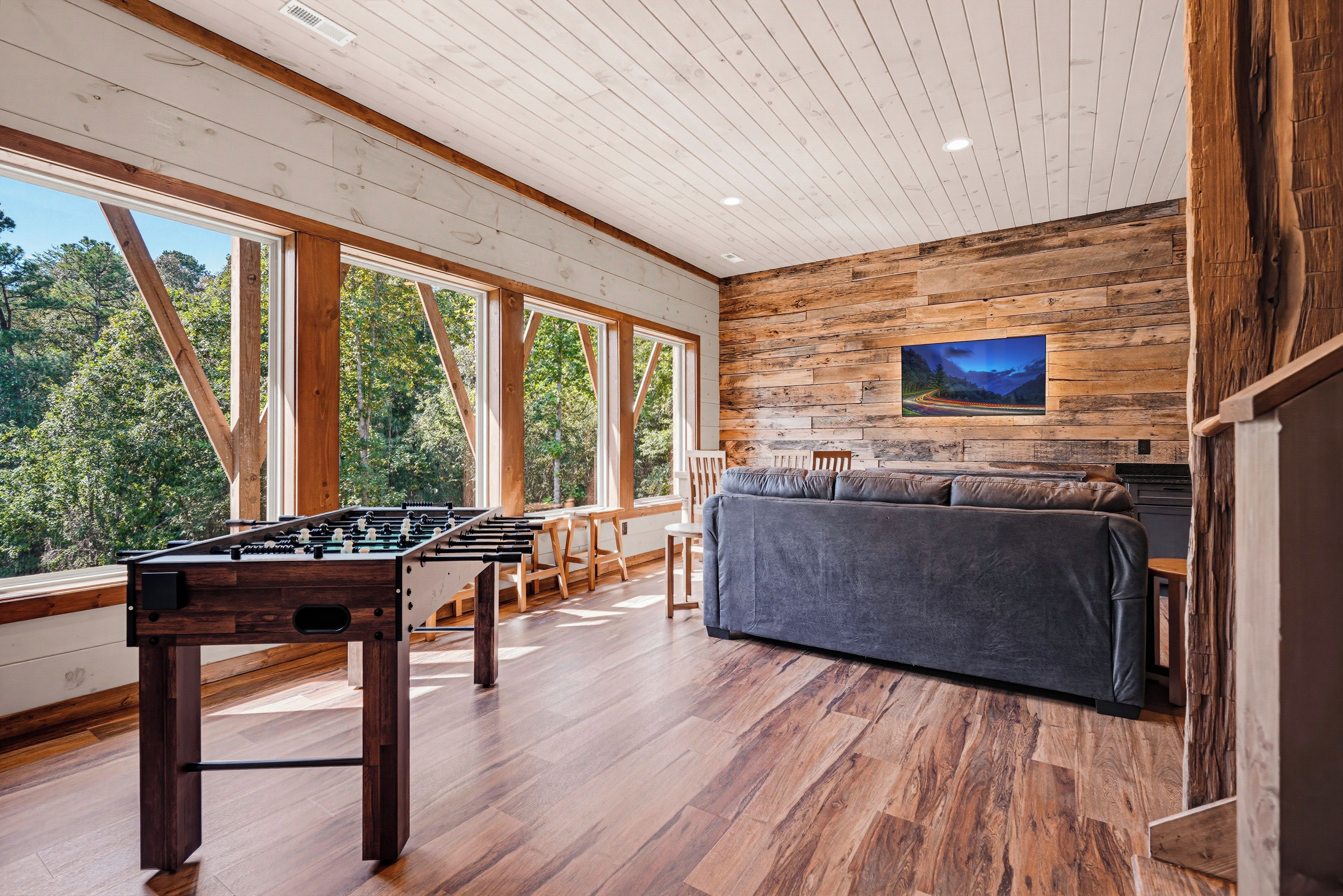 Una habitación amplia con mesa de fútbol, sofá y un ventanal que ofrece vistas a árboles.