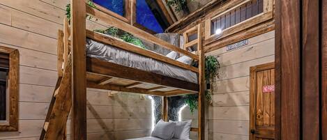 Ein Schlafraum mit Etagenbetten und Blick auf einen sternenübersäten Himmel durch ein Fenster, mit Holzwänden und einer Tür mit der Aufschrift 'TREEHOUSE'.