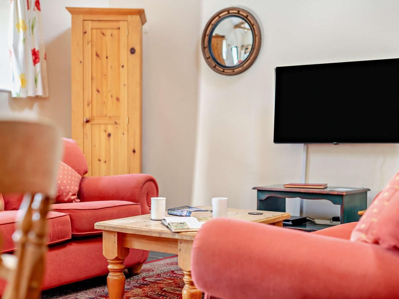 A living room with a red sofa, a wooden cabinet, a round mirror, a television, and a small table with books and a cup.