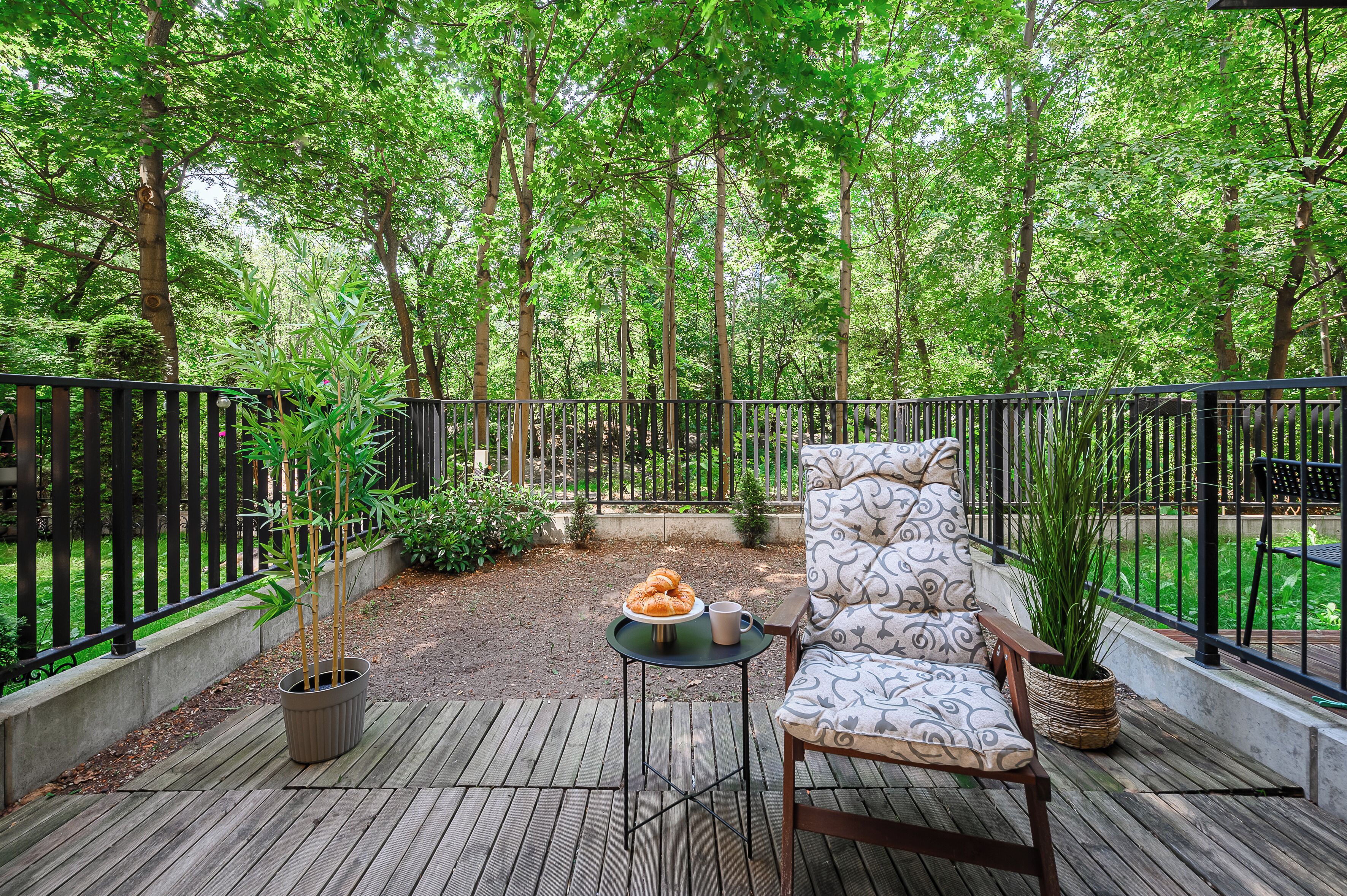 A wooden deck with a chair and table, surrounded by a metal fence, overlooking a lush green forest.