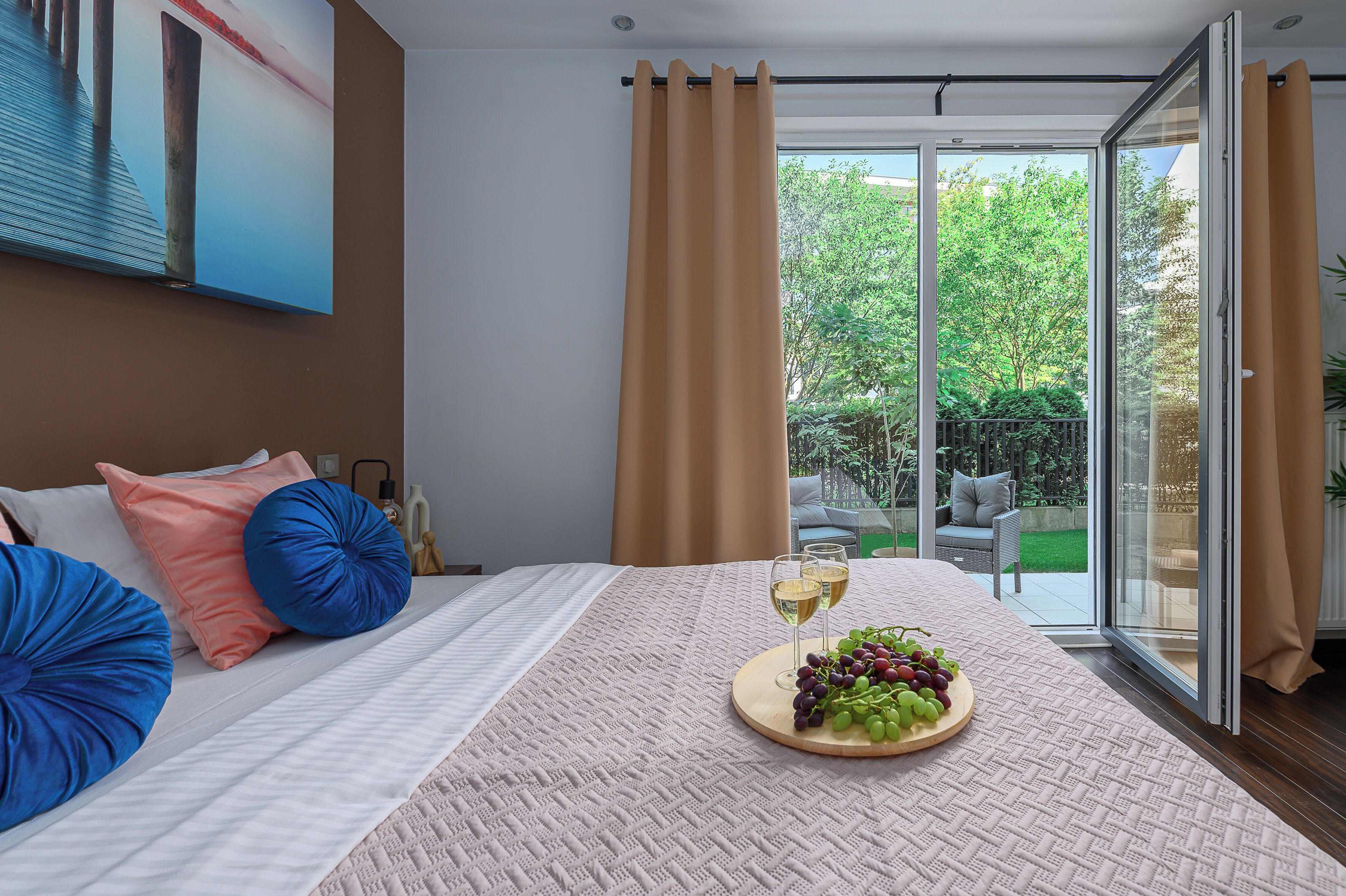 A bedroom with a bed, a glass door leading to a balcony, and a table with a fruit platter and glasses.