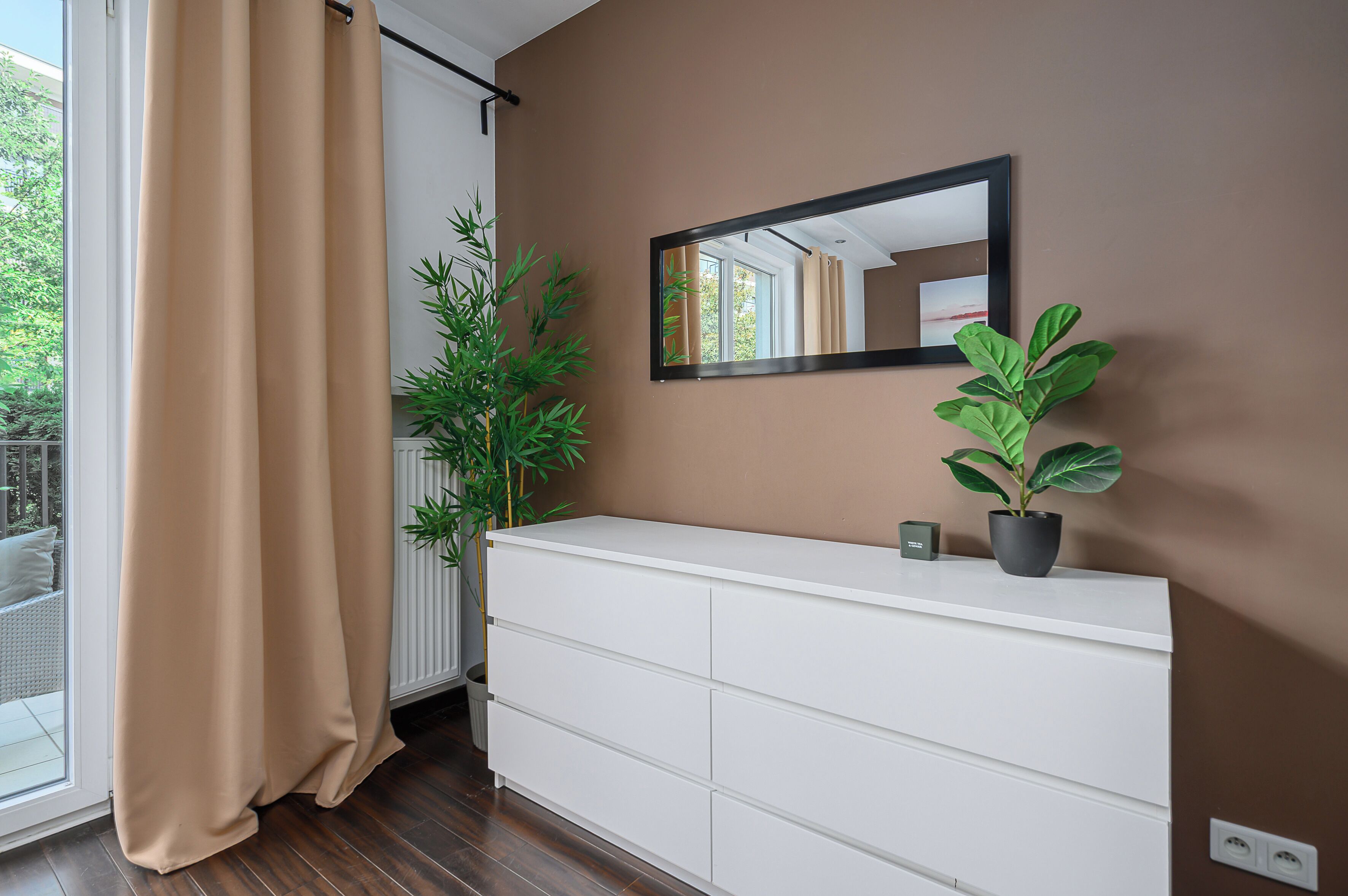 Ein Zimmer mit weißem Schminktisch, einem Spiegel, einer Pflanze und einem Fenster mit Vorhängen.