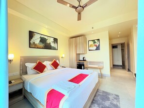 Une chambre d’hôtel comprenant un lit, des tables de chevet, un ventilateur de plafond, des œuvres d’art accrochées aux murs et une porte donnant sur une autre pièce.