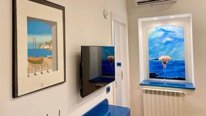 Una habitación con un cuadro enmarcado de una escena costera, un televisor de pantalla plana y un ventanal con una pintura marina.