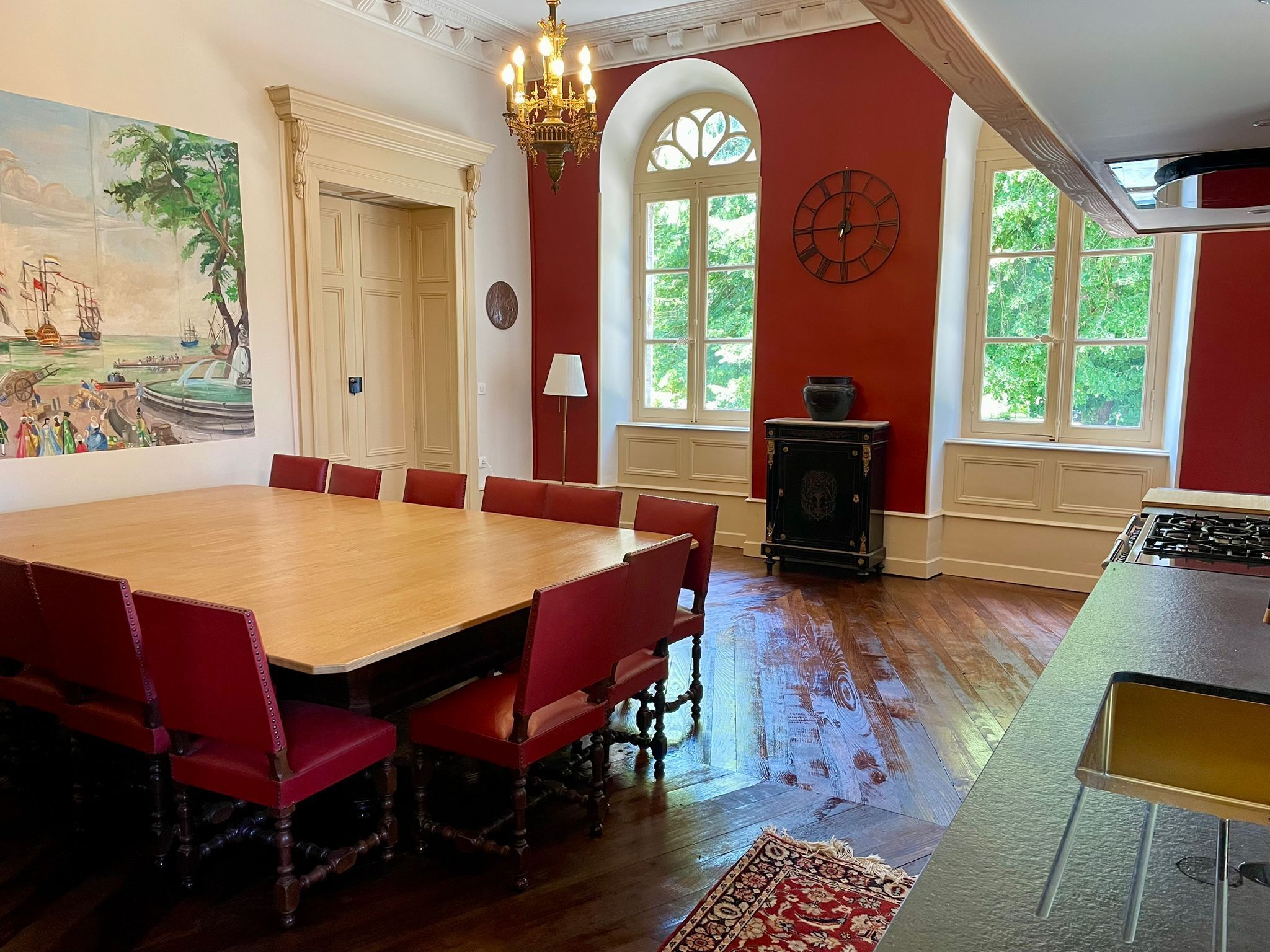 A dining room with a large wooden table, red upholstered chairs, a chandelier, and a painting on the wall.