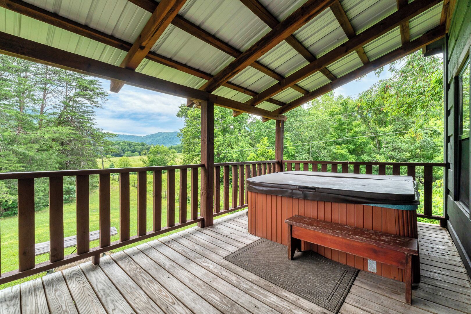 A wooden deck with a hot tub and bench, surrounded by a metal roof and wooden railings, overlooking a green landscape.