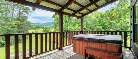 A wooden deck with a hot tub and bench, surrounded by a metal roof and wooden railings, overlooking a green landscape.