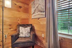 Habitación de cabaña de madera con una silla acolchada, una lámpara de pared y un ventanal con persianas.