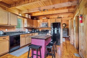 Ein Holzblockhaus-Interieur mit einer Küche, die eine zentrale Kochinsel, Barhocker, eine Mikrowelle, einen Kühlschrank und einen Feuerlöscher umfasst.