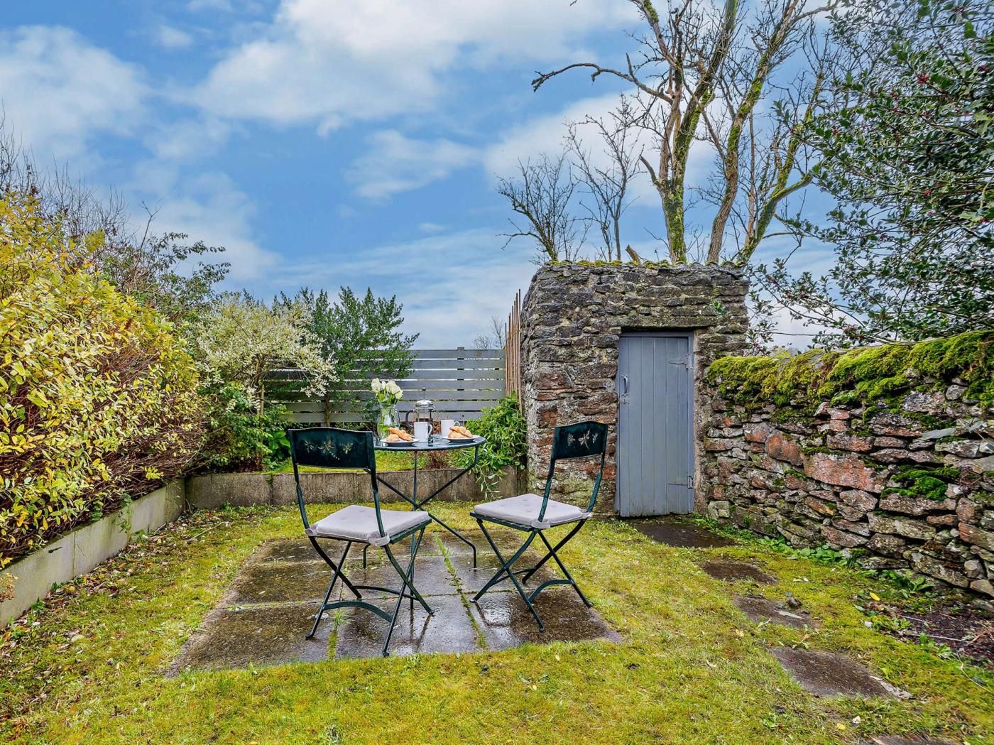 Un mur de pierre, une petite porte et une table avec des chaises dans un jardin.
