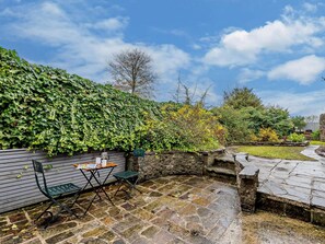 A patio area with a stone floor, a wooden table with a chair, and a green hedge. - 1 Bed in Monyash (oc-pk760) (Monyash)