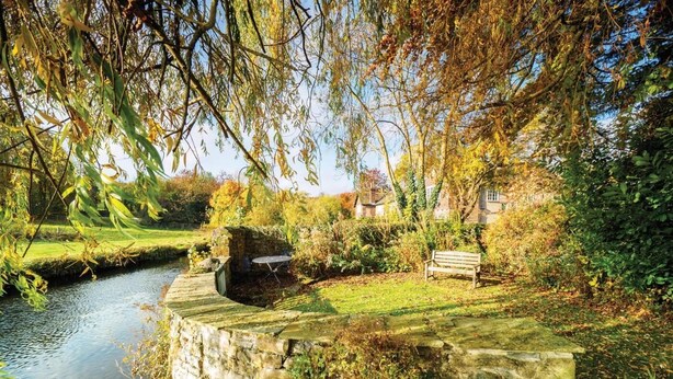 Eine steinerne Brücke über einem ruhigen Fluss, umgeben von üppiger Vegetation und einer Bank.