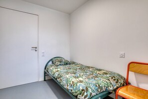 Ein kleines Zimmer mit einem Einzelbett, einer Tür, einer Steckdose und einem Stuhl.