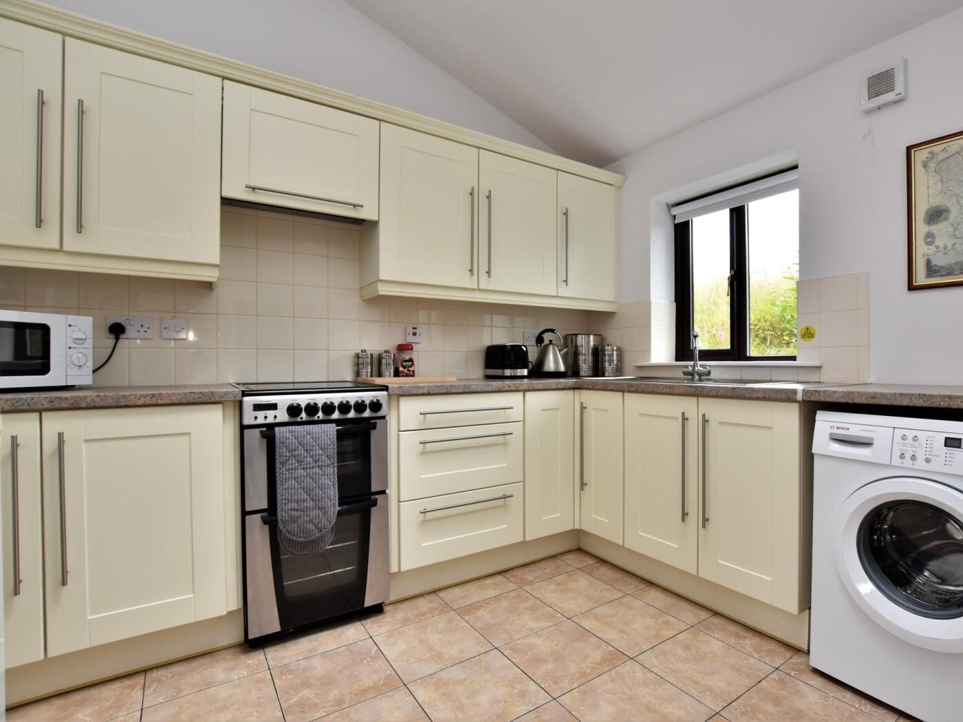 A kitchen with cream-colored cabinets, a microwave, an oven, a washing machine, and a window.