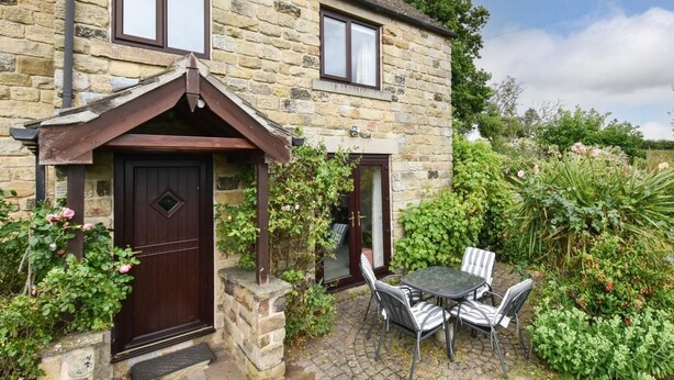 A stone house with a dark brown door and windows, a small garden with a table and chairs, and a cobblestone pathway. - 2 Bed in Holmesfield (oc-pk783) (Dronfield)