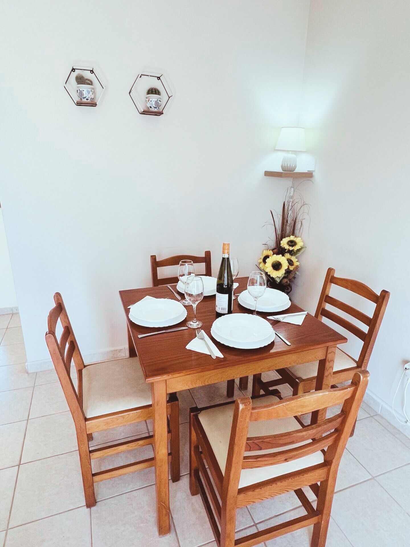 Mesa de comedor lista para cuatro personas, con sillas de madera, una botella de vino y un jarrón de girasoles.