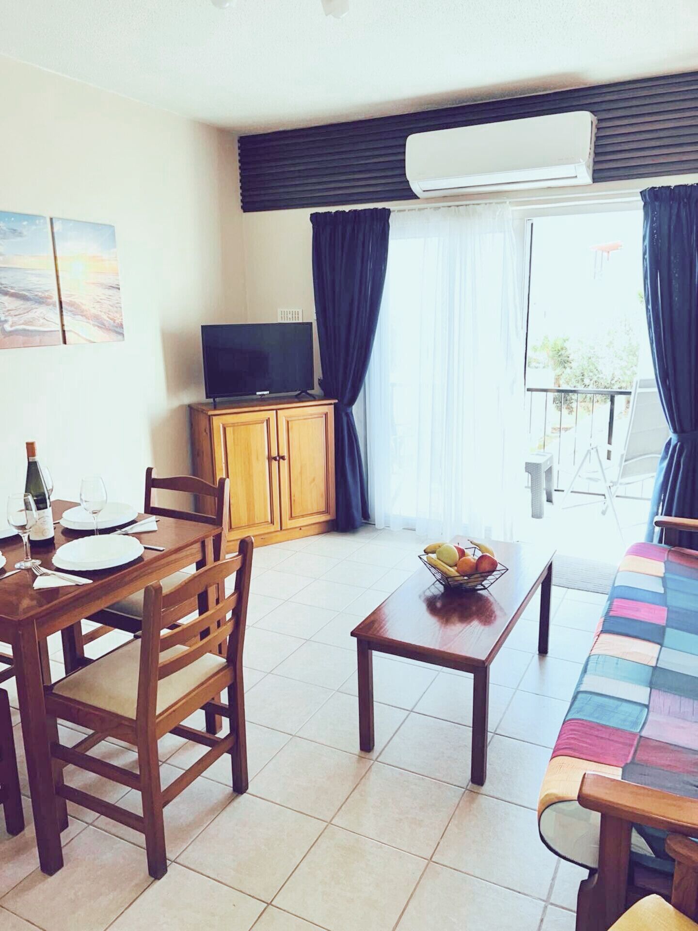 Habitación de hotel con comedor, televisor sobre un mueble de madera y balcón con cortinas.