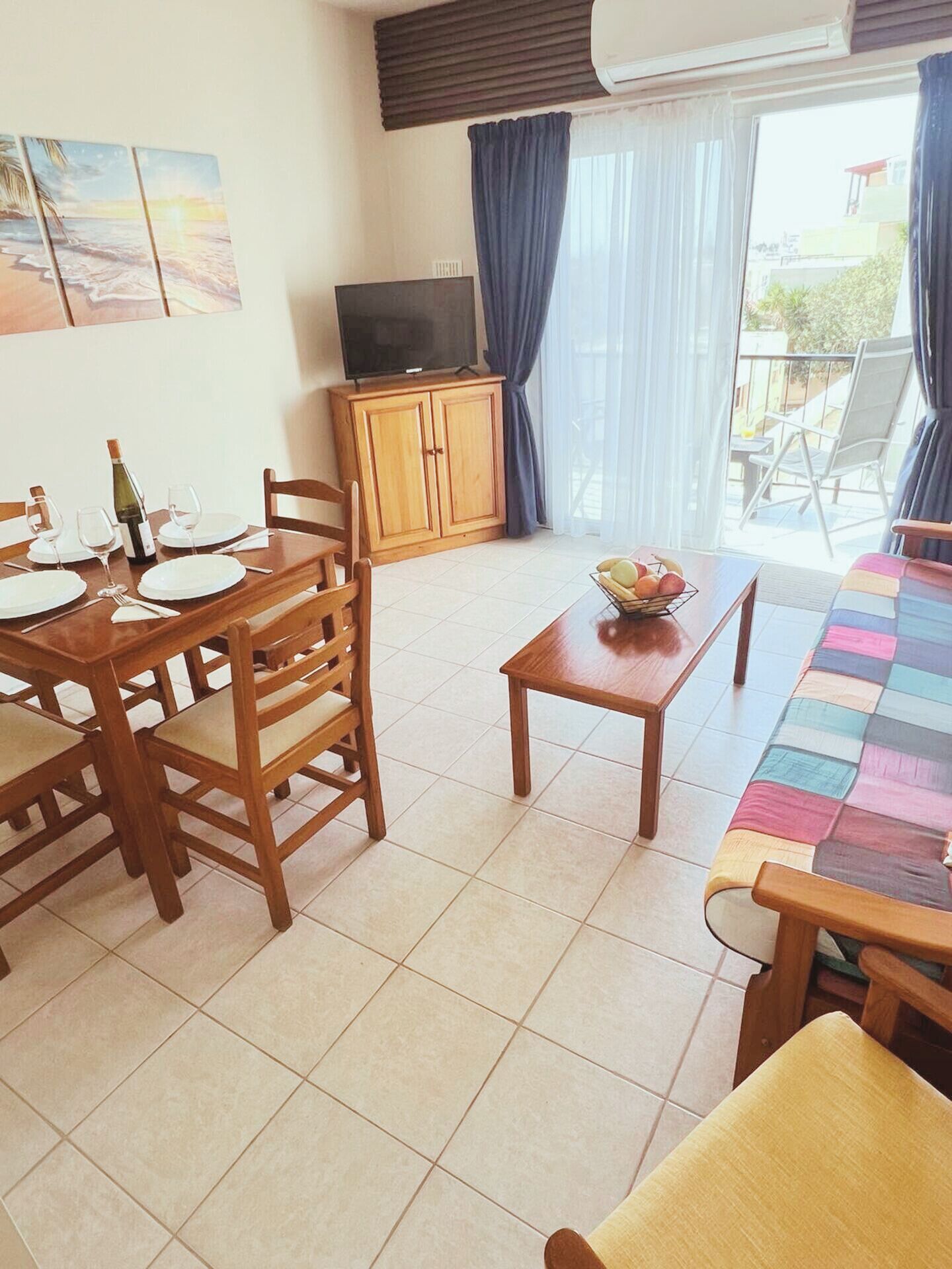 Un comedor con una mesa lista para cuatro, un televisor sobre un mueble, un armario y un balcón con sillas.
