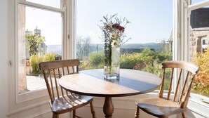 A round wooden table with two chairs, a vase with flowers, and a view of greenery and buildings through large windows. - 4 Bed in Matlock (oc-pk861) (Holloway)
