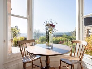 Ein runder Holztisch mit zwei Stühlen, eine Vase mit Blumen und Blick auf Grünflächen und Gebäude durch große Fenster.