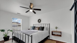 Un dormitorio con cama, ventilador de techo, ventana con persianas, una planta en maceta y una pequeña estantería.