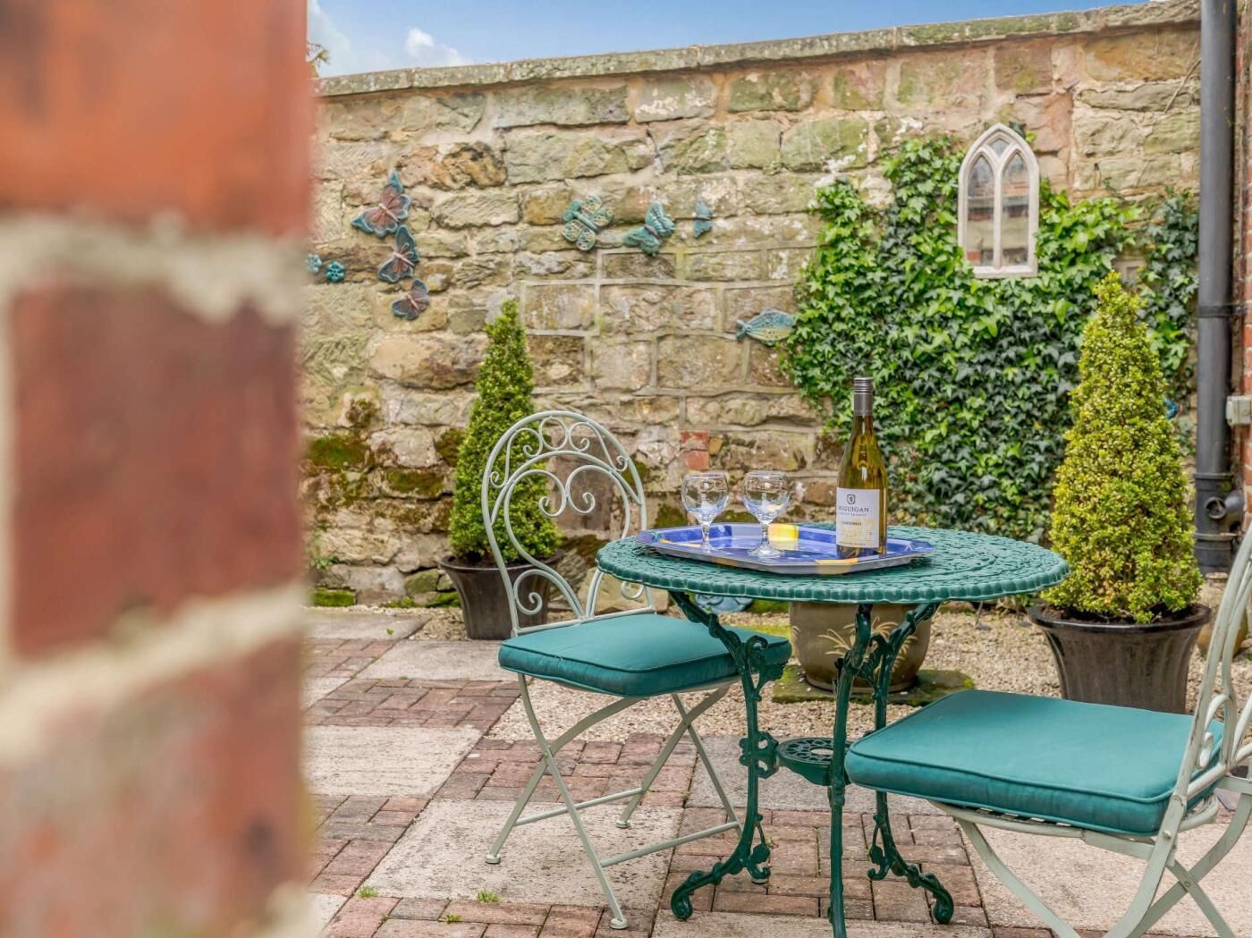 A stone wall with ivy, a round table with chairs, and a bottle of wine.