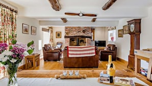 Un salón acogedor con chimenea, sofás de cuero, una mesa de madera con una botella y aperitivos, y un reloj en la pared.