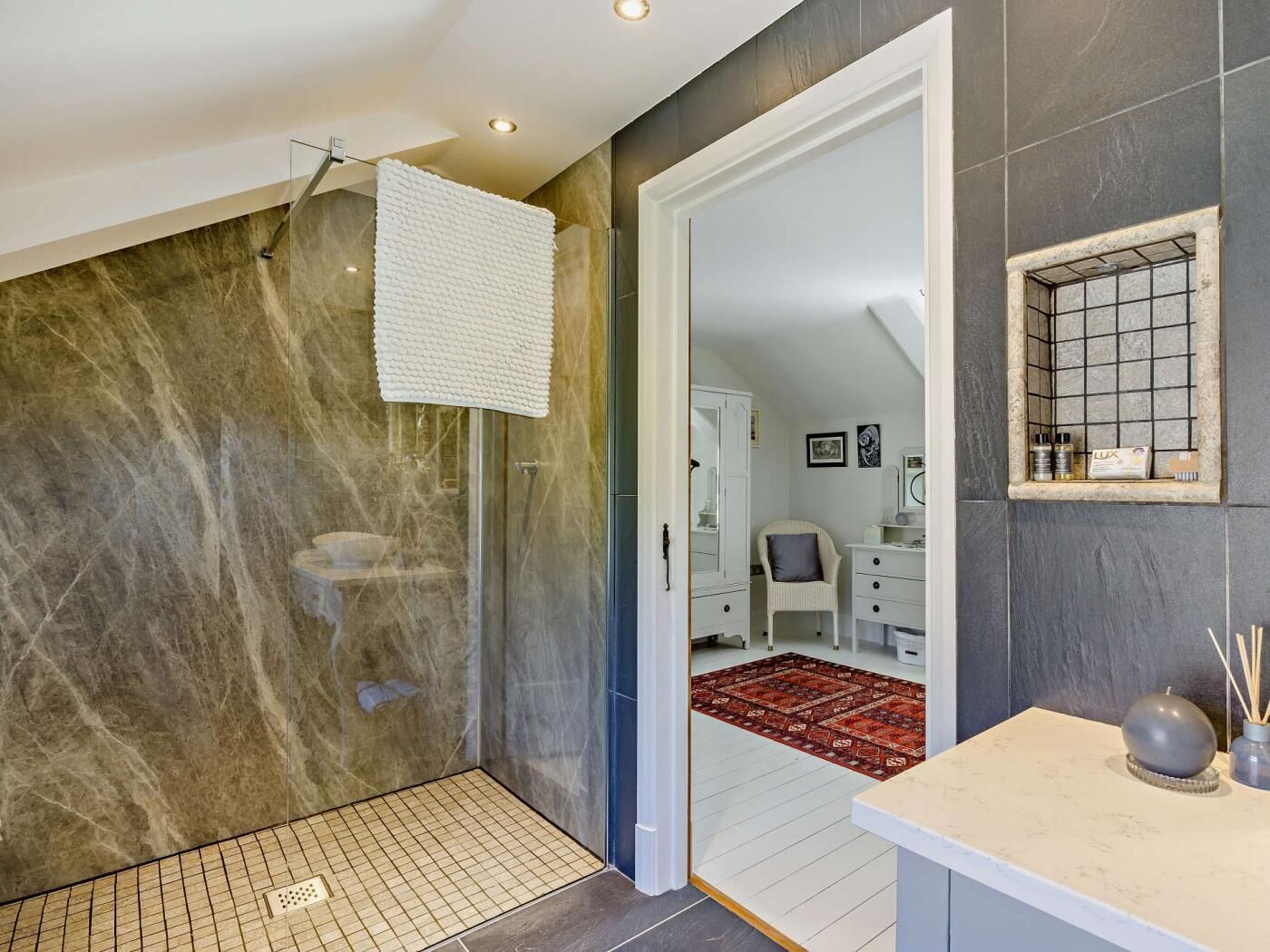 Une salle de bain moderne avec une douche à parois de verre, une serviette blanche accrochée au mur, un meuble-lavabo blanc avec un miroir et un tapis rouge à motifs sur le sol.