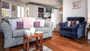 A living room with a grey sofa, a blue armchair, wooden flooring, and a kitchen in the background. - 3 Bed in Baslow (oc-pk917) (Baslow)