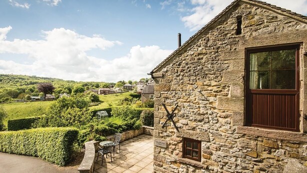 A stone building with a large window, a door, and a small window. There is a paved patio with chairs and a table. - 1 Bed in Wirksworth (oc-pk925) (Wirksworth)