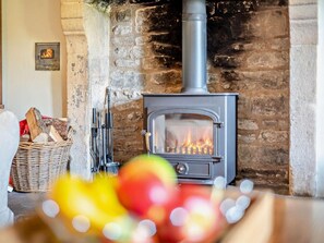 A stone fireplace with a modern wood-burning stove, a basket of firewood, and a blurred foreground of colorful objects. - 4 Bed in Hope Valley (oc-pk939) (Calver)