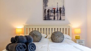 Een net opgemaakt bed met opgerolde handdoeken, twee decoratieve kussens, een schilderij van een regenachtig straatbeeld en twee bedlampen.