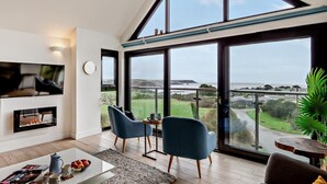 A spacious room with a large bay window, a flat-screen TV, a fireplace, two blue chairs, a coffee table, and a view of the sea. - 2 Bed in Helston (oc-45739) (Kuggar)