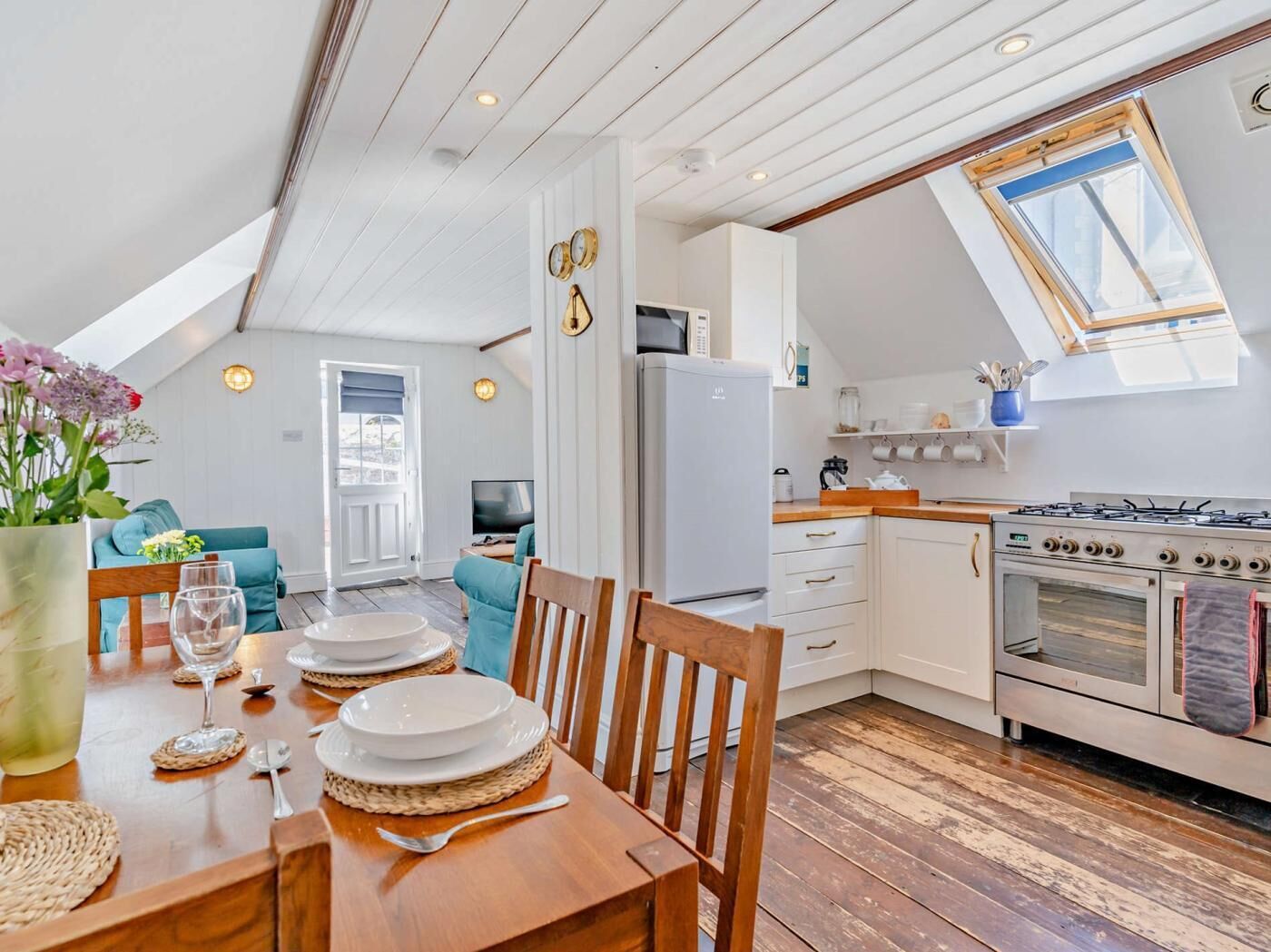 A modern kitchen with a gas stove, oven, and white cabinets. There is a dining area with a wooden table set for two, and a living room with a sofa and a TV.
