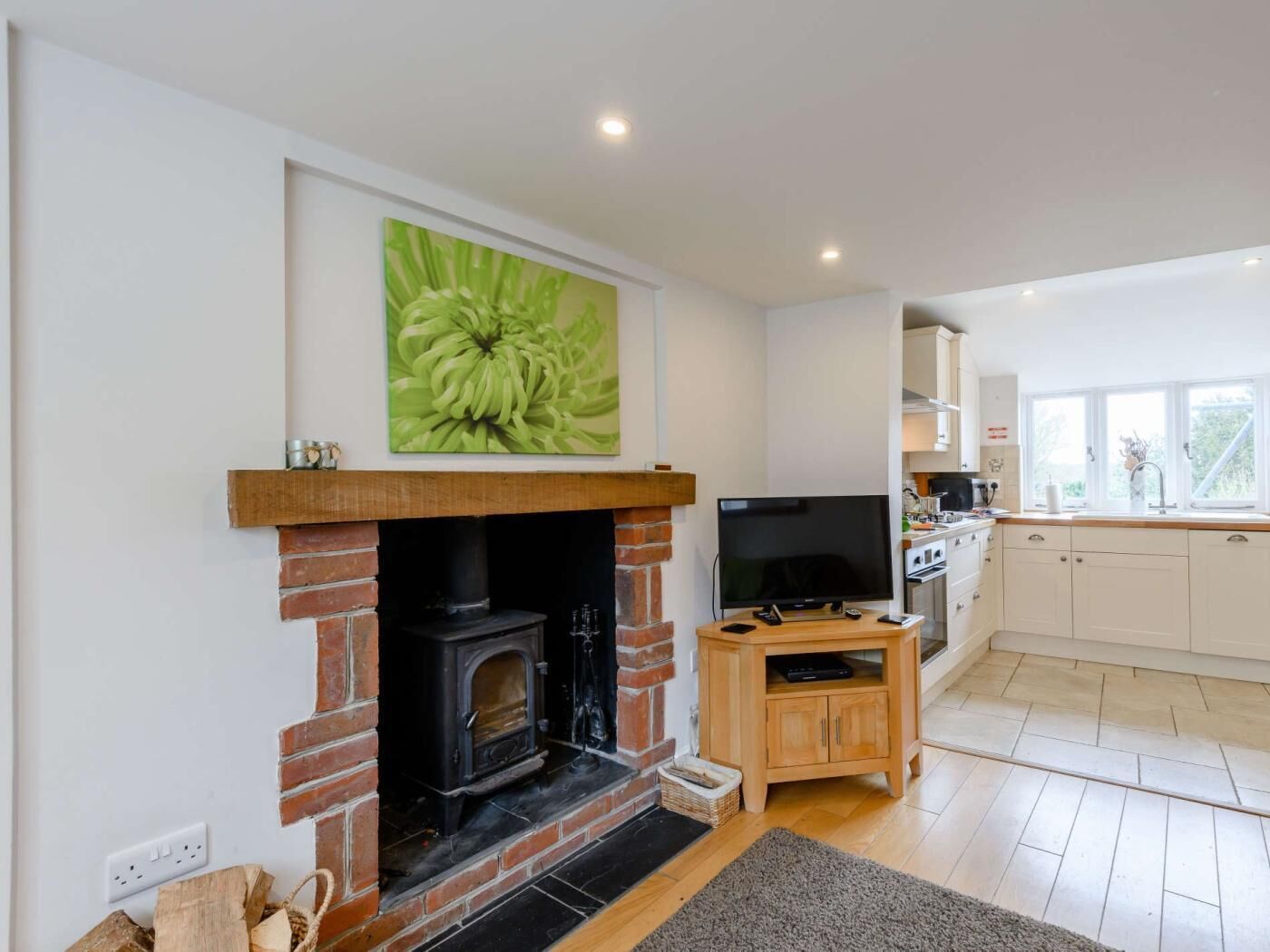 A living room with a fireplace, a television, and a kitchen area with white cabinets and a window.