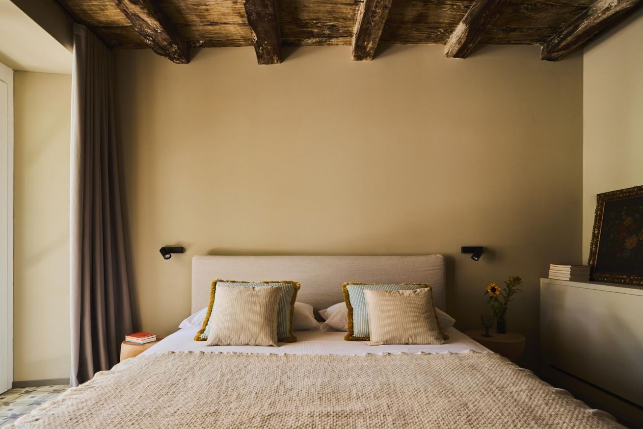Une chambre avec un plafond en bois, un lit doté d’une tête de lit beige, deux oreillers, une table de chevet avec un vase de fleurs et un tableau encadré accroché au mur.