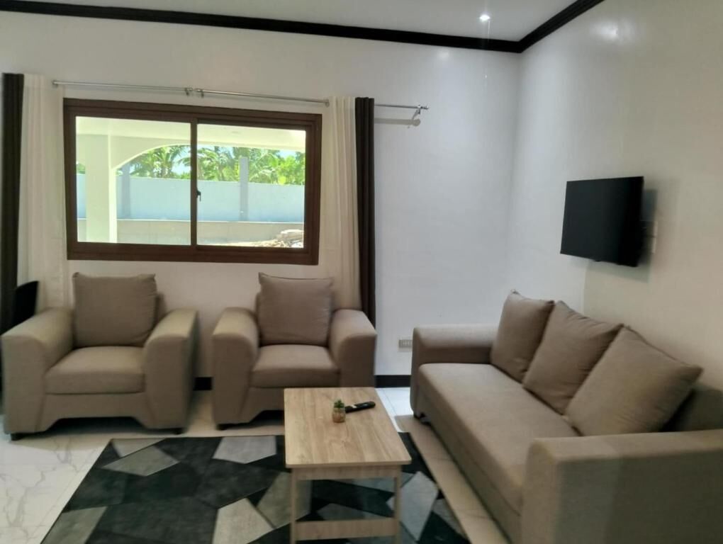 Ein Wohnzimmer mit zwei Sofas, einem Couchtisch, einem an der Wand montierten Fernseher und einem großen Fenster mit Vorhängen.