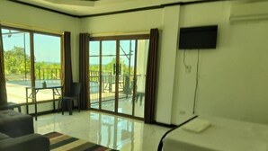 A modern hotel room with a large sliding glass door leading to a balcony, a sofa, a TV mounted on the wall, and a clean, shiny floor.