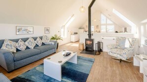 A spacious loft with a high ceiling, a built-in wood stove, a sofa, a coffee table, and a patterned armchair. - 3 Bed in Bideford (oc-46252) (Northam)