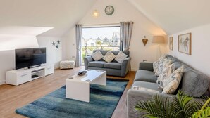 A modern living room with a flat-screen TV, a sofa, a coffee table, and a large window with curtains. - 3 Bed in Bideford (oc-46252) (Northam)