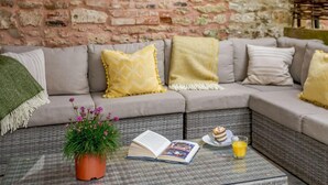 A cozy seating area with a brick wall, a sofa with cushions, a small table with a book, a potted plant, and a glass of orange juice. - 4 Bed in Honiton (oc-45563) (Payhembury)
