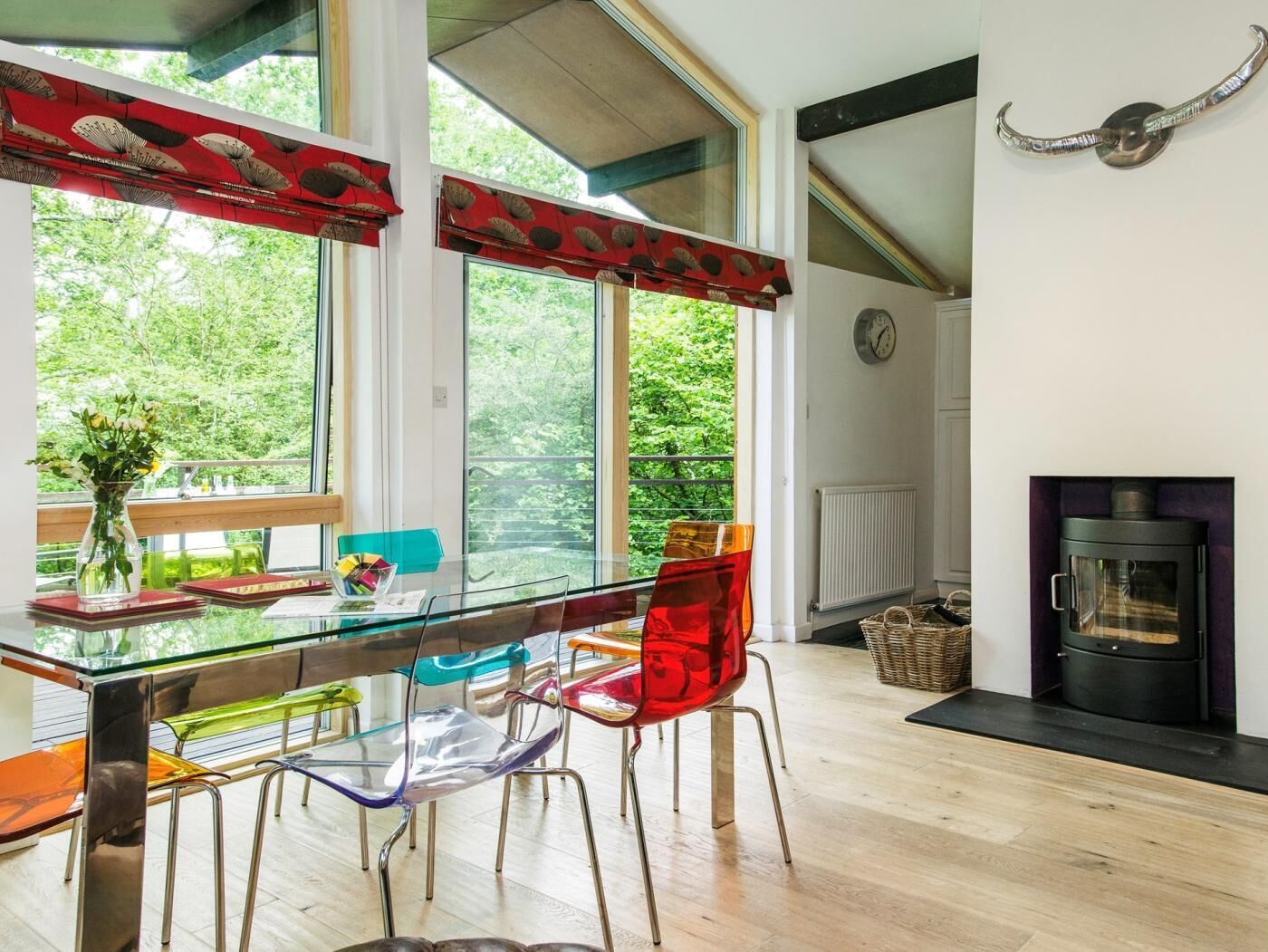 A modern dining area with a glass table, colorful chairs, a view of greenery outside, and a fireplace.