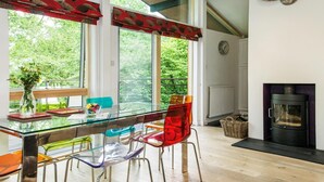 A modern dining area with a glass table, colorful chairs, a view of greenery outside, and a fireplace. - 2 Bed in Beaulieu (oc-nfl71) (Beaulieu)