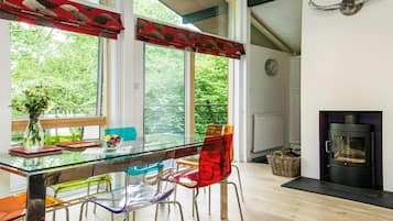 A modern dining area with a glass table, colorful chairs, a view of greenery outside, and a fireplace.