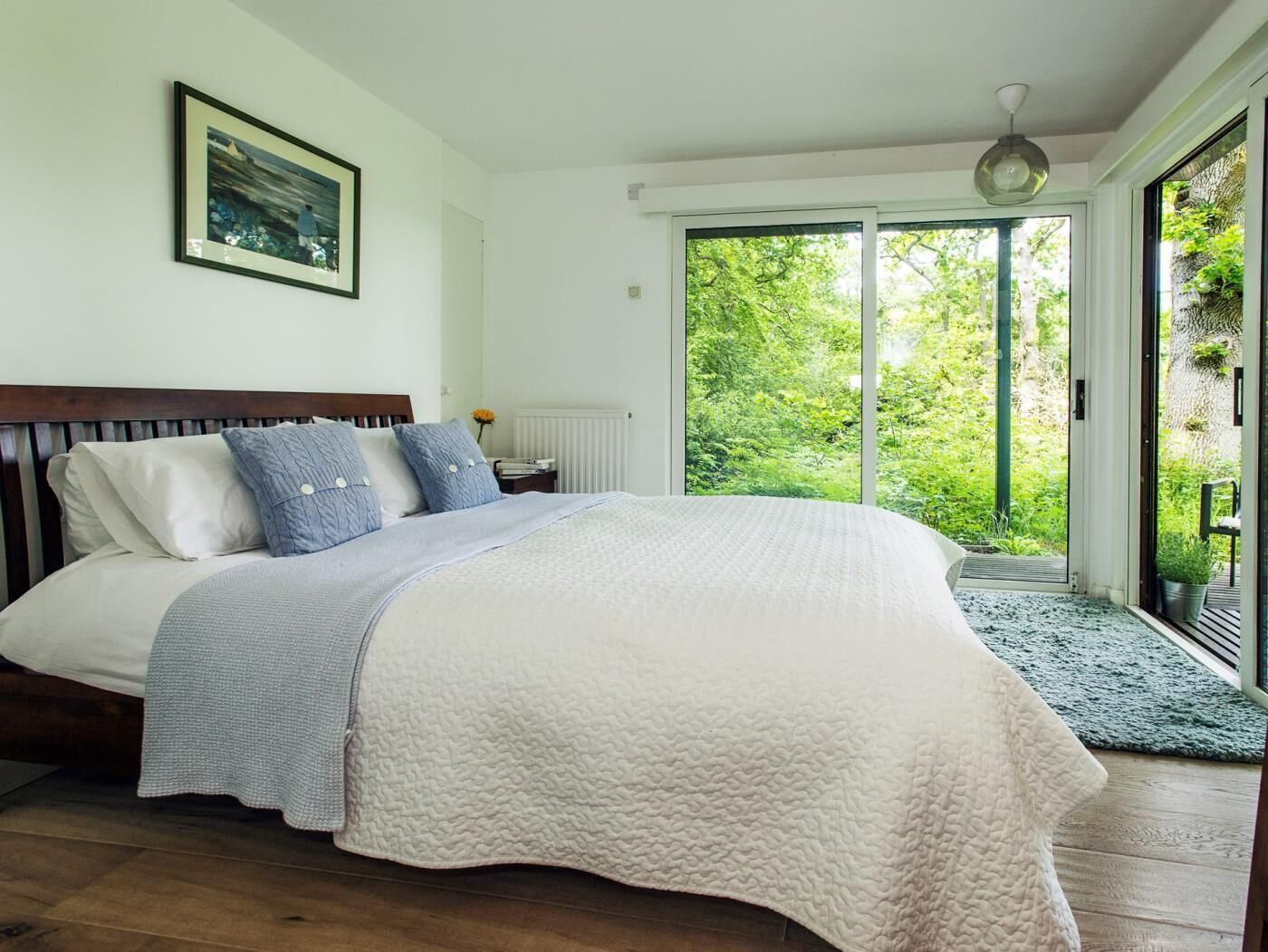 A bedroom with a large bed, a wooden headboard, a framed picture on the wall, and a view of greenery through a sliding glass door.